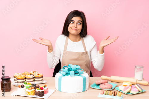 Pastry chef with a big cake in a table over isolated pink background making doubts gesture
