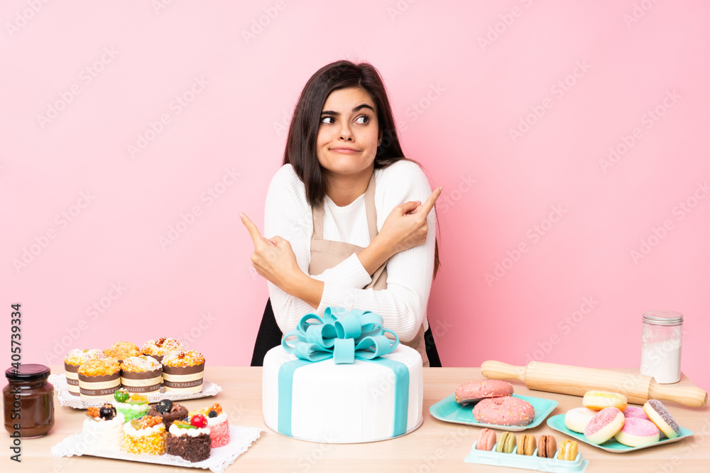 Pastry chef with a big cake in a table over isolated pink background pointing to the laterals having doubts
