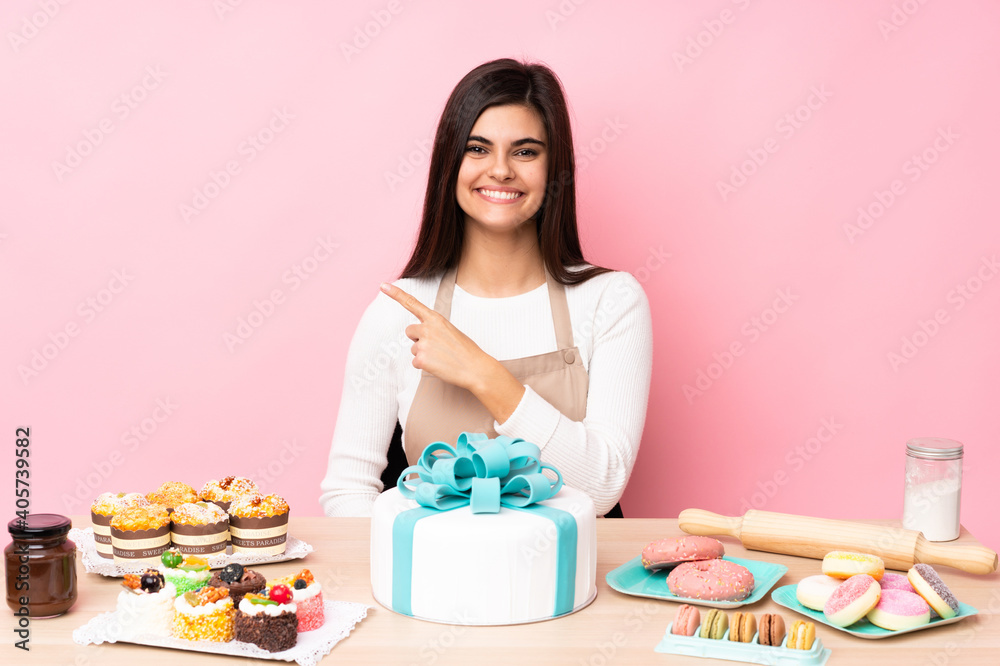 Pastry chef with a big cake in a table over isolated pink background pointing to the side to present a product Pastry chef with a big cake in a table over isolated pink background pointing to the side to present a product