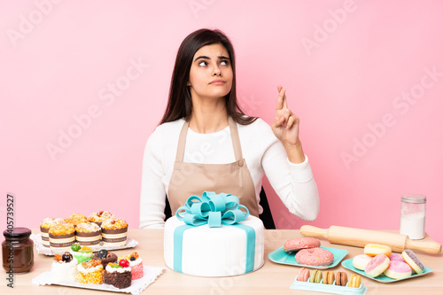 Pastry chef with a big cake in a table over isolated pink background with fingers crossing and wishing the best