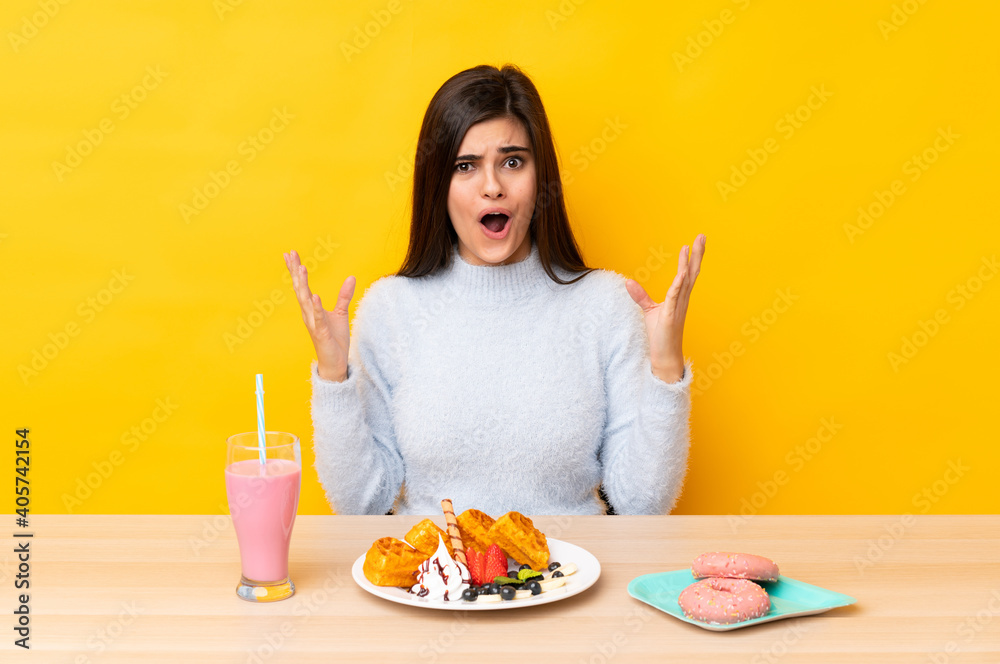 Young woman eating waffles and milkshake in a table over isolated yellow background with surprise facial expression Young woman eating waffles and milkshake in a table over isolated yellow background with surprise facial expression