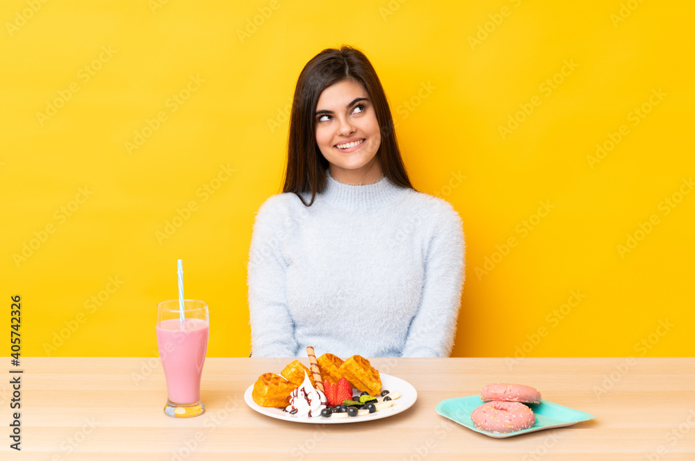 Young woman eating waffles and milkshake in a table over isolated yellow background laughing and looking up Young woman eating waffles and milkshake in a table over isolated yellow background laughing and looking up