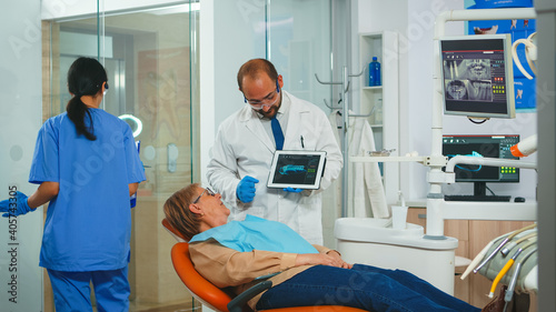 Dentistry doctor showing x-ray of teeth to patient using tablet in dental clinic. Stomatologist reviewing dental radiography with senior woman explaining treatment sitting on stomatological chair.