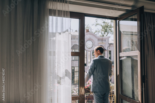 Stylish groom, man, gentleman stands near the window on the terrace, veranda in the hotel against the backdrop of the city on a sunny day. Wedding portrait.