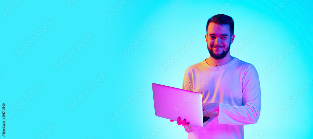 Using laptop. Caucasian man's portrait with device on blue studio background in neon light. Beautiful male model. Concept of human emotions, facial expression, sales, ad. Copyspace for ad, flyer Using laptop. Caucasian man's portrait with device on blue studio background in neon light. Beautiful male model. Concept of human emotions, facial expression, sales, ad. Copyspace for ad, flyer