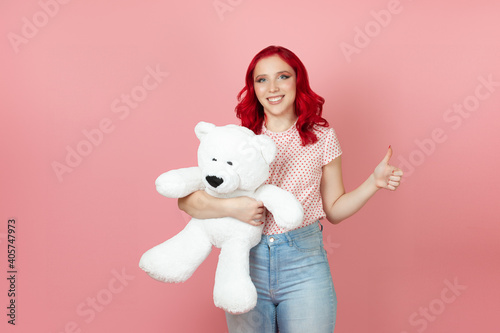 a smiling, delighted young woman with red hair holds a large white teddy bear and gives a thumbs up isolated on a pink background.