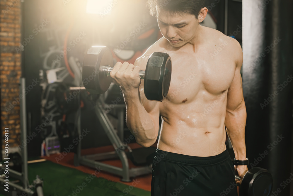 A handsome and shapely Asian man lifting dumbbells and looking up at the arm muscles in a fitness gym. A handsome and shapely Asian man lifting dumbbells and looking up at the arm muscles in a fitness gym.