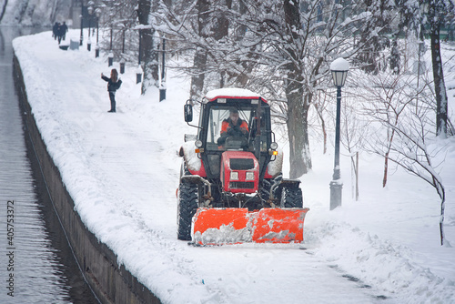 Snow removal tractor with rotating brush sweeping snow from footpath on embankment in park. Red tractor with scoop and automated brush removes snow. Municipal road sweeping vehicle with plow