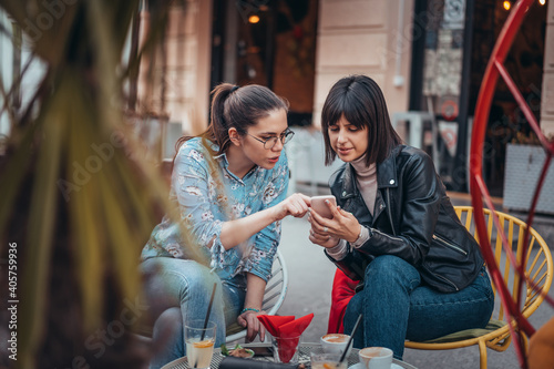Two beautiful women in cafe outdoors looking at something on mobile phone. Two female using mobile phone application shopping online and enjoying coffee outside in city