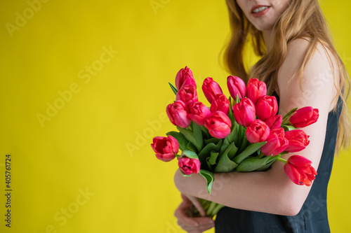 Unrecognizable woman with a bouquet of red tulips on a yellow background. A girl in a black dress holds an armful of flowers. A gift for International Women's Day. Spring holiday.