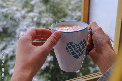 person drinking hot chocolate with pink marshmallows in a mug with blue heart across the winter landscape in open window. Winter vacation. St. Valentine's day photo