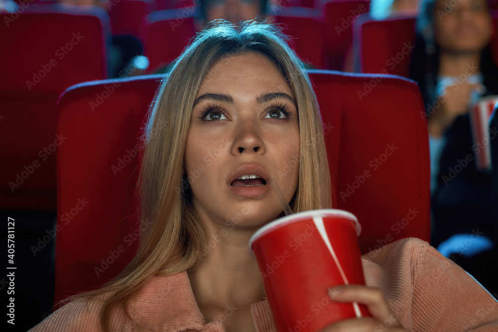 Portrait of pretty young woman looking excited and having a drink while watching movie alone in cinema auditorium