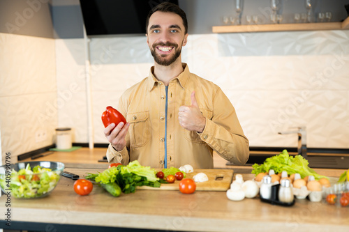 Handsome guy with beard and nice smile is giving a thumb on healthy lifestyle and eating fresh food. Young vegetarian man is cooking salad in kitchen indoor.