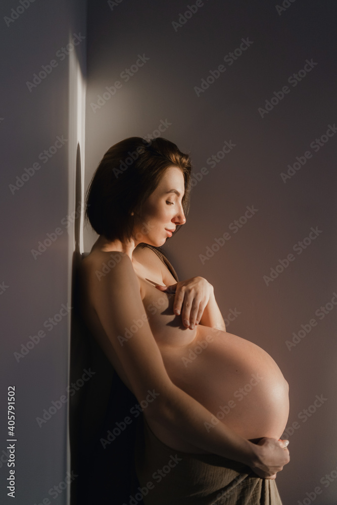 Young pregnant girl without clothes in the rays of the sun in the bedroom by the wall Young pregnant girl without clothes in the rays of the sun in the bedroom by the wall