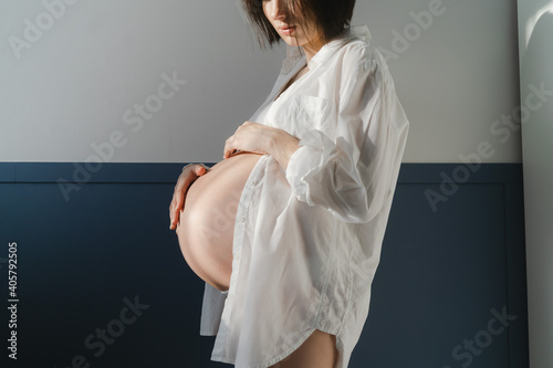 Young pregnant girl in white underwear and white shirt near a white wall in the sun