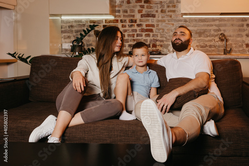 Dad with a beard, son, and young mom with long hair are watching TV and smiling on the sofa. The family is enjoying a happy evening at home. A child is listening to his mother.