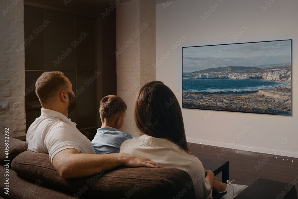 A photo from behind of a dad with a beard, a son, and a young mom which are watching a movie on a widescreen television set on the sofa. The family is enjoying a video together in the evening at home.