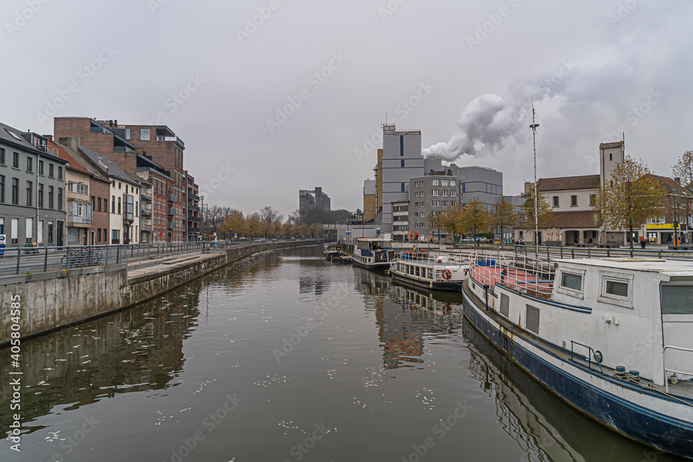 Beautifull picture of the river the dender in Aalst on a foggy day with boats in the background and autumn trees in orange red.  River with road under bridge. East flanders, belgium on foggy day