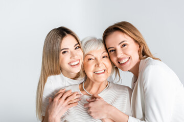 three generation of positive women smiling while looking at camera and hugging isolated on grey