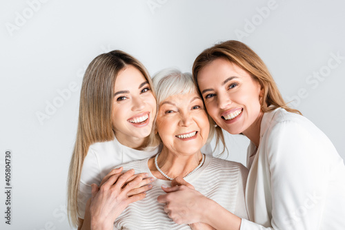 three generation of positive women smiling while looking at camera and hugging isolated on grey photo