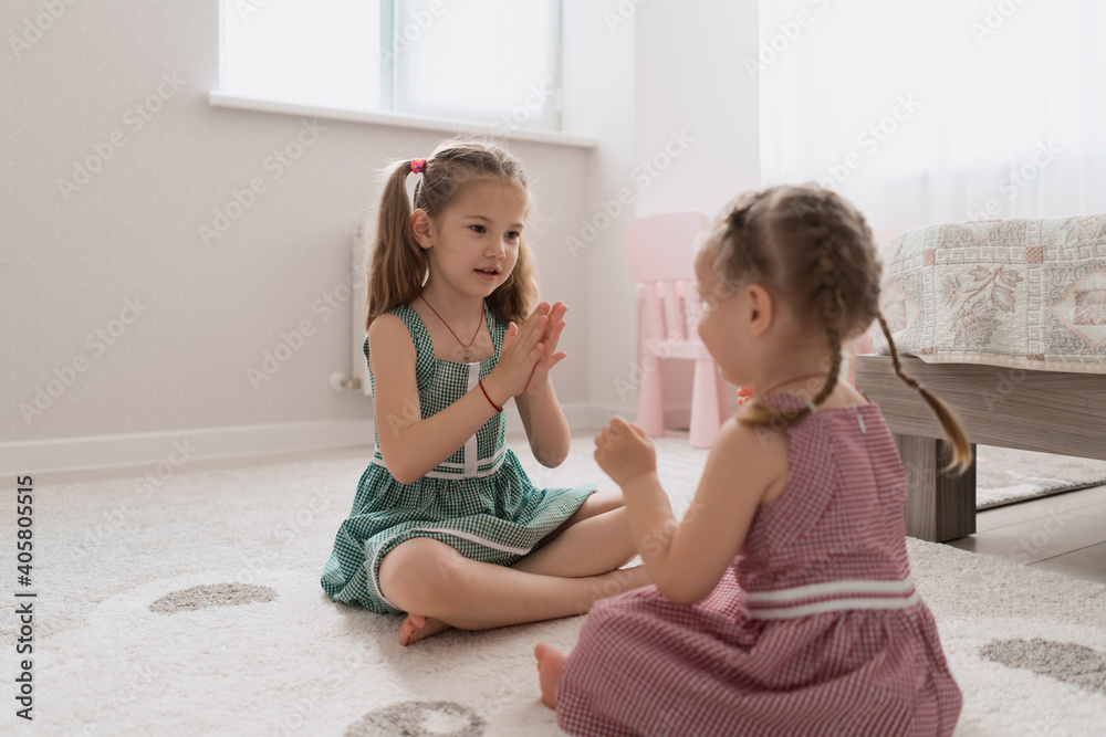 Cute little sisters playing together sitting on the carpet in identical dresses different colors in a room with lots of light Cute little sisters playing together sitting on the carpet in identical dresses different colors in a room with lots of light