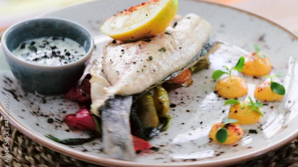 Close up slow motion view sea bass served with bbq vegetables, sauce and slice of lemon on spinning plate. Restaurant main course dish concept