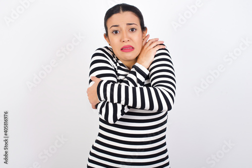 Desperate Young beautiful woman wearing stripped t-shirt against white background trembles and feels cold, hugs oneself to warm up or feels scared notices something terrifying.