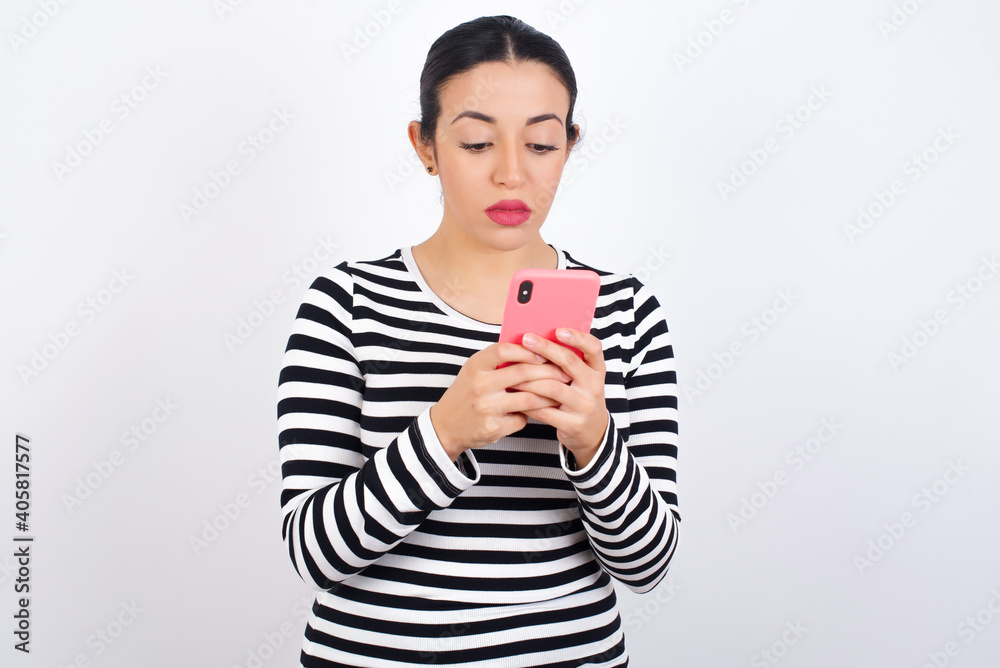 Excited Young beautiful woman wearing stripped t-shirt against white background winking and eye hold smart phone use read social network news Excited Young beautiful woman wearing stripped t-shirt against white background winking and eye hold smart phone use read social network news
