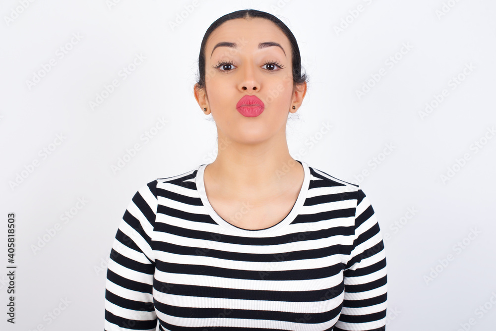Shot of pleasant looking Young beautiful woman wearing stripped t-shirt against white background, pouts lips, looks at camera, Human facial expressions Shot of pleasant looking Young beautiful woman wearing stripped t-shirt against white background, pouts lips, looks at camera, Human facial expressions