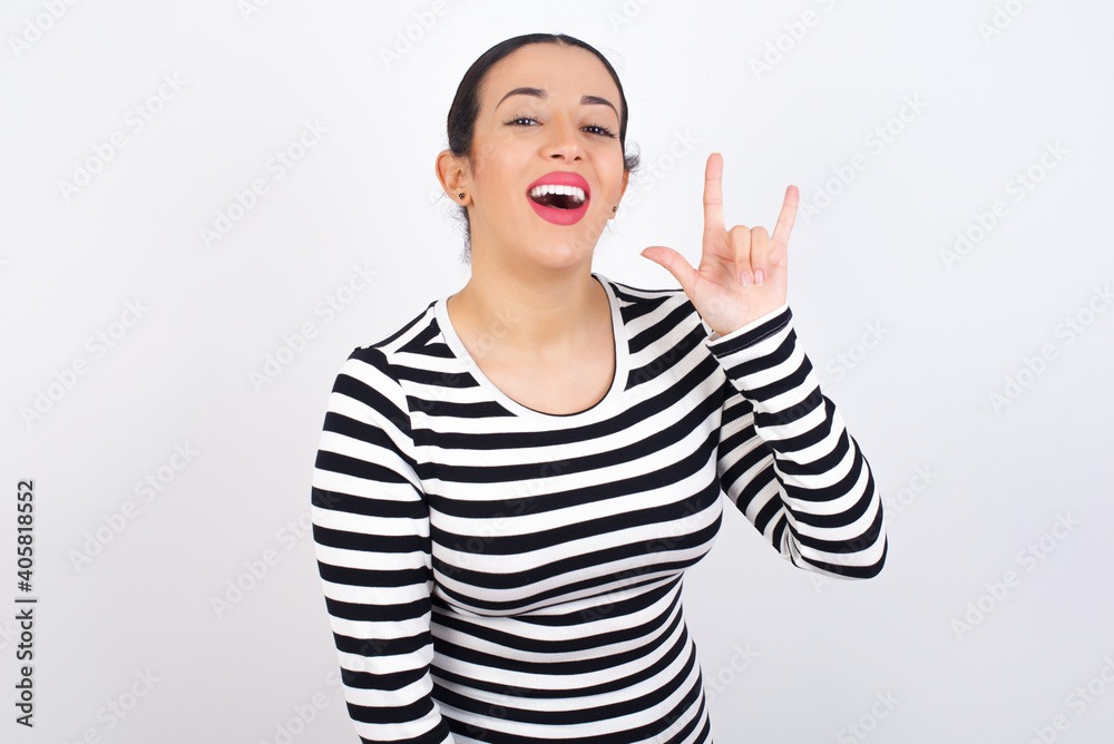 Young beautiful woman wearing stripped t-shirt against white background doing a rock gesture and smiling to the camera. Ready to go to her favorite band concert. Young beautiful woman wearing stripped t-shirt against white background doing a rock gesture and smiling to the camera. Ready to go to her favorite band concert.