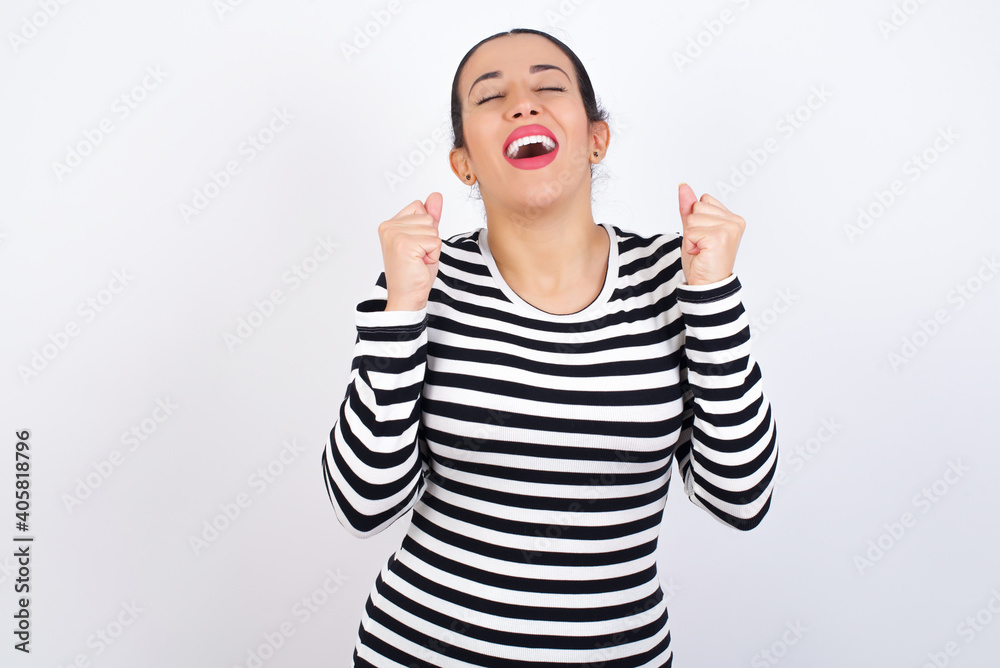 Young beautiful woman wearing stripped t-shirt against white background being excited for success with raised arms and closed eyes celebrating victory. Winner concept. Young beautiful woman wearing stripped t-shirt against white background being excited for success with raised arms and closed eyes celebrating victory. Winner concept.