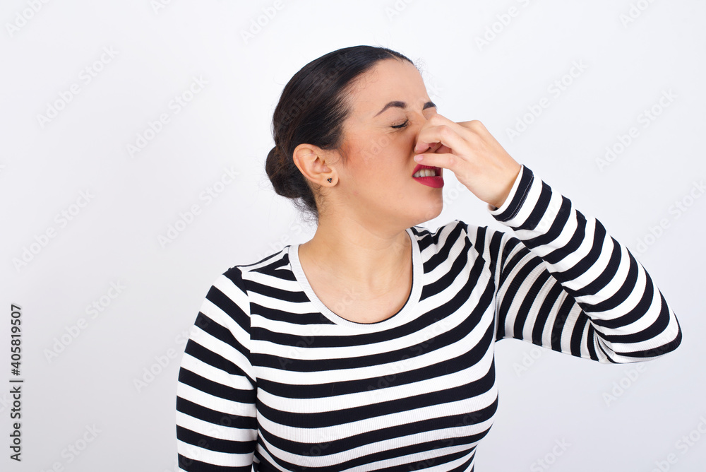 Young beautiful woman wearing stripped t-shirt against white background smelling something stinky and disgusting, intolerable smell, holding breath with fingers on nose. Bad smell Young beautiful woman wearing stripped t-shirt against white background smelling something stinky and disgusting, intolerable smell, holding breath with fingers on nose. Bad smell