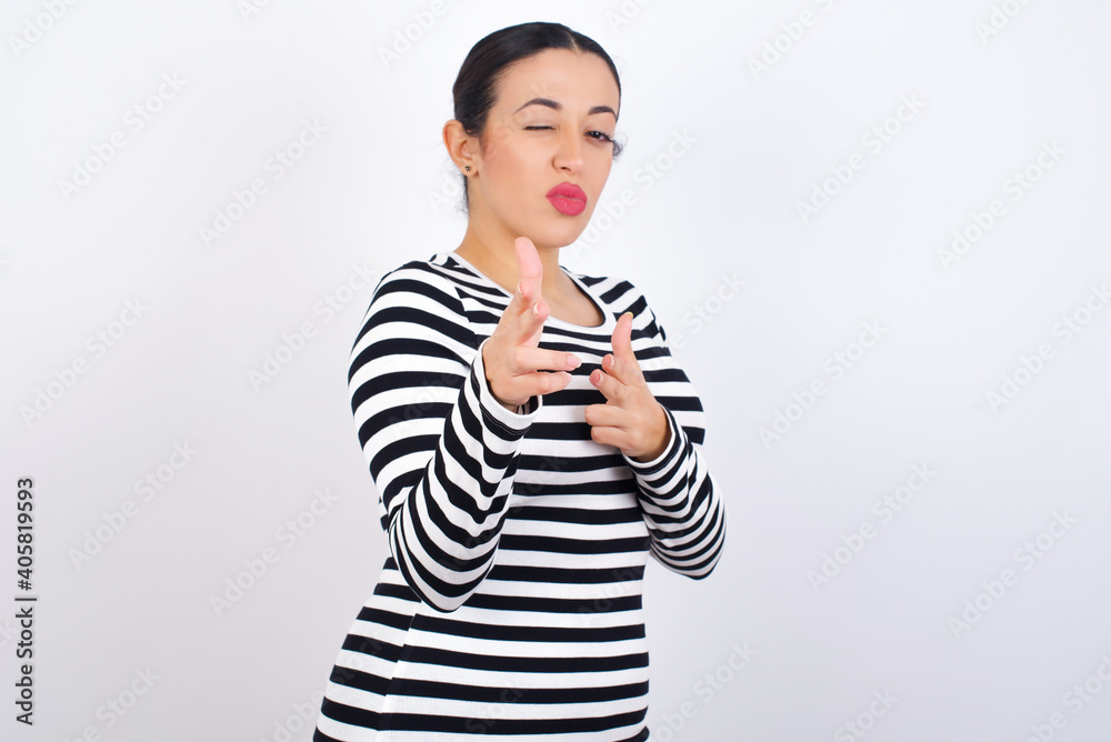 Joyful Young beautiful woman wearing stripped t-shirt against white background wink and points index fingers at camera, chooses someone, makes finger gun pistol. Joyful Young beautiful woman wearing stripped t-shirt against white background wink and points index fingers at camera, chooses someone, makes finger gun pistol.