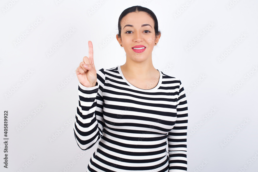 Young beautiful woman wearing stripped t-shirt against white background showing and pointing up with finger number one while smiling confident and happy. Young beautiful woman wearing stripped t-shirt against white background showing and pointing up with finger number one while smiling confident and happy.