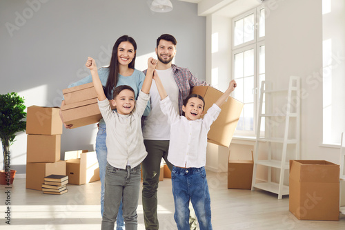 Happy family with little children celebrating moving into new home. Portrait of smiling young couple with excited kids standing in room with packed boxes. Buying house, real estate, mortgage concept