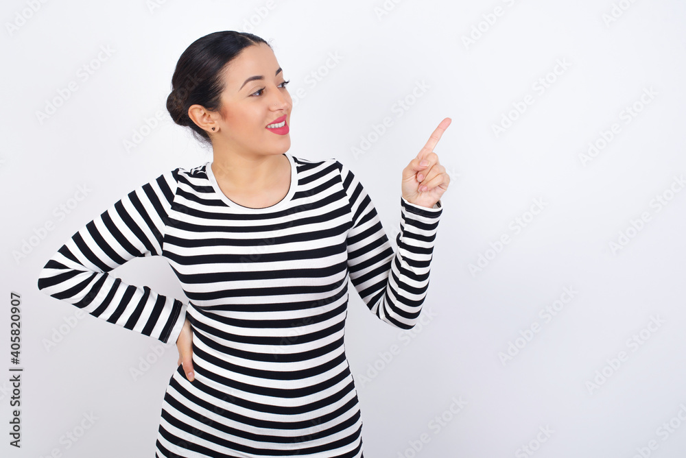 Smiling Young beautiful woman wearing stripped t-shirt against white background indicating finger empty space showing best low prices Smiling Young beautiful woman wearing stripped t-shirt against white background indicating finger empty space showing best low prices