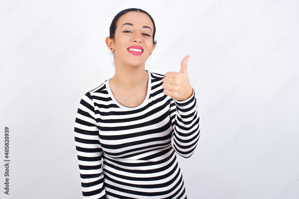 Young beautiful woman wearing stripped t-shirt against white background doing happy thumbs up gesture with hand. Approving expression looking at the camera showing success. Young beautiful woman wearing stripped t-shirt against white background doing happy thumbs up gesture with hand. Approving expression looking at the camera showing success.