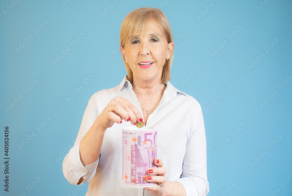 Senior woman puts money into the piggy bank over blue background Senior woman puts money into the piggy bank over blue background