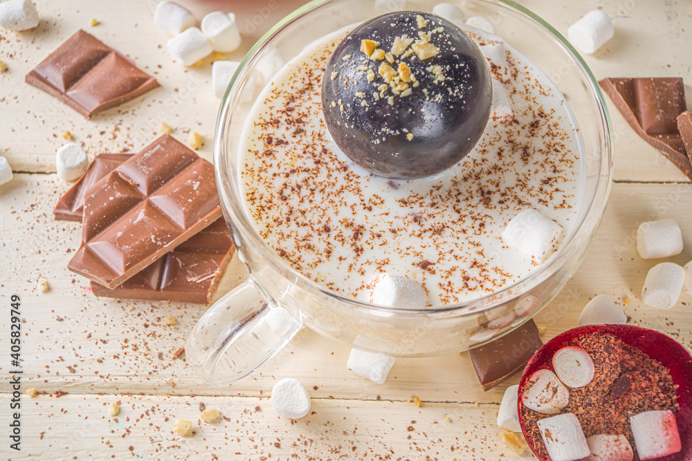 Hot chocolate bomb, with marshmallow and chocolates and nuts,  Woman's hand dropping chocolate bomb into cup with milk. - obrazy, fototapety, plakaty 