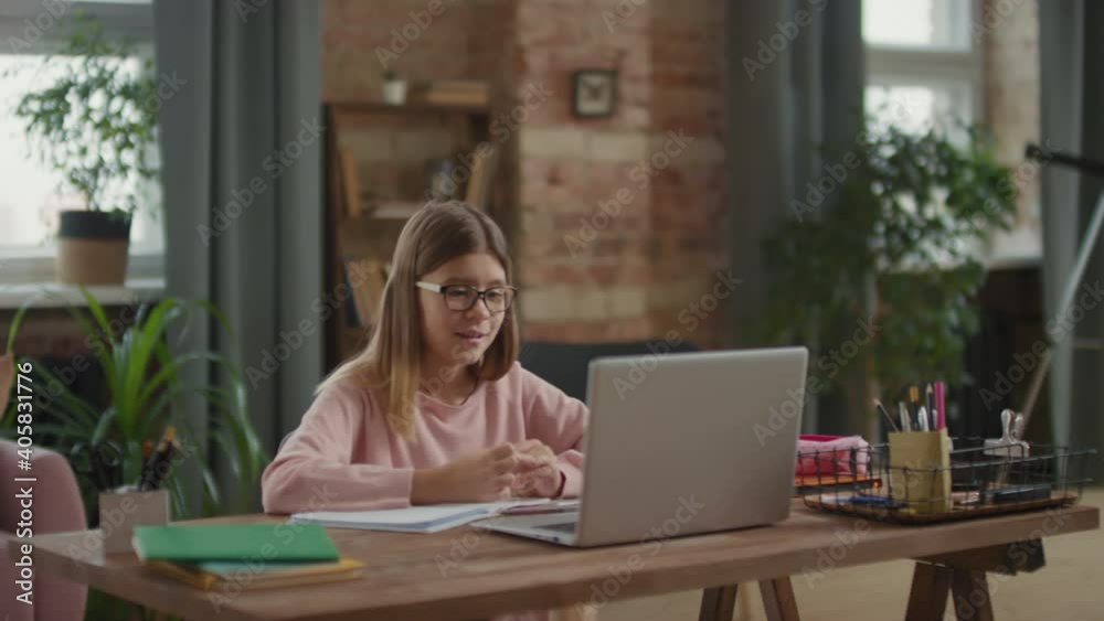 Medium shot of young girl sitting at desk studying online via laptop while caring woman playing with kid on sofa in background Medium shot of young girl sitting at desk studying online via laptop while caring woman playing with kid on sofa in background