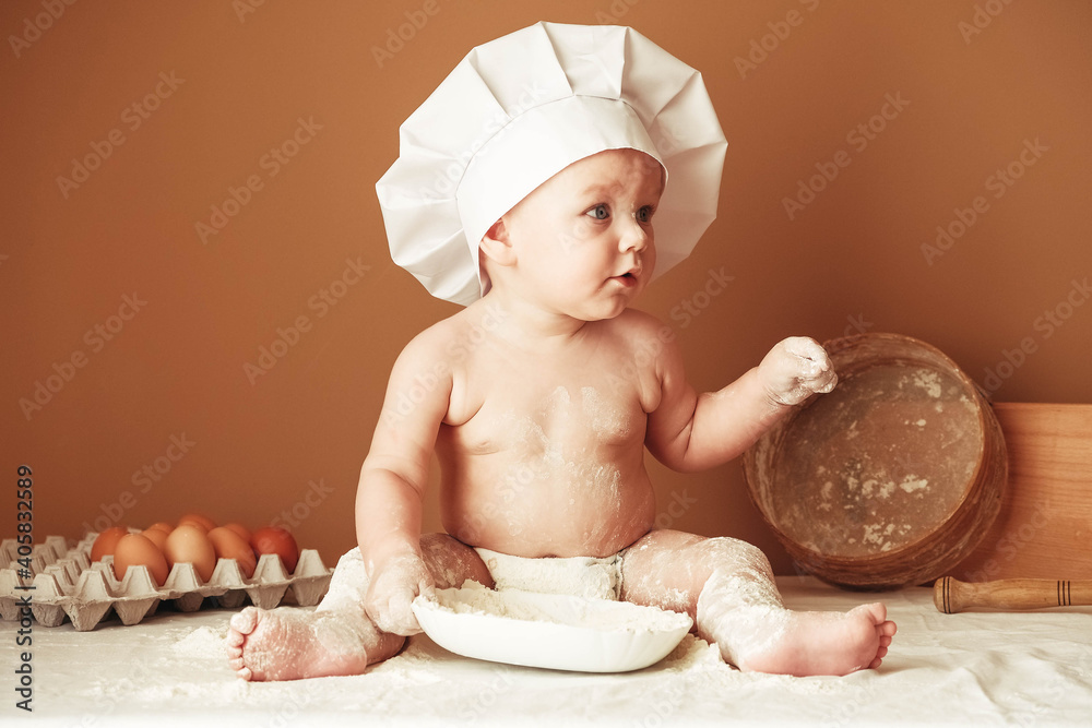 Little boy baker in a chef's hat sitting on the table playing with flour on a brown background with a wooden rolling pin, a round rustic sieve and eggs. Copy, empty space for text Little boy baker in a chef's hat sitting on the table playing with flour on a brown background with a wooden rolling pin, a round rustic sieve and eggs. Copy, empty space for text