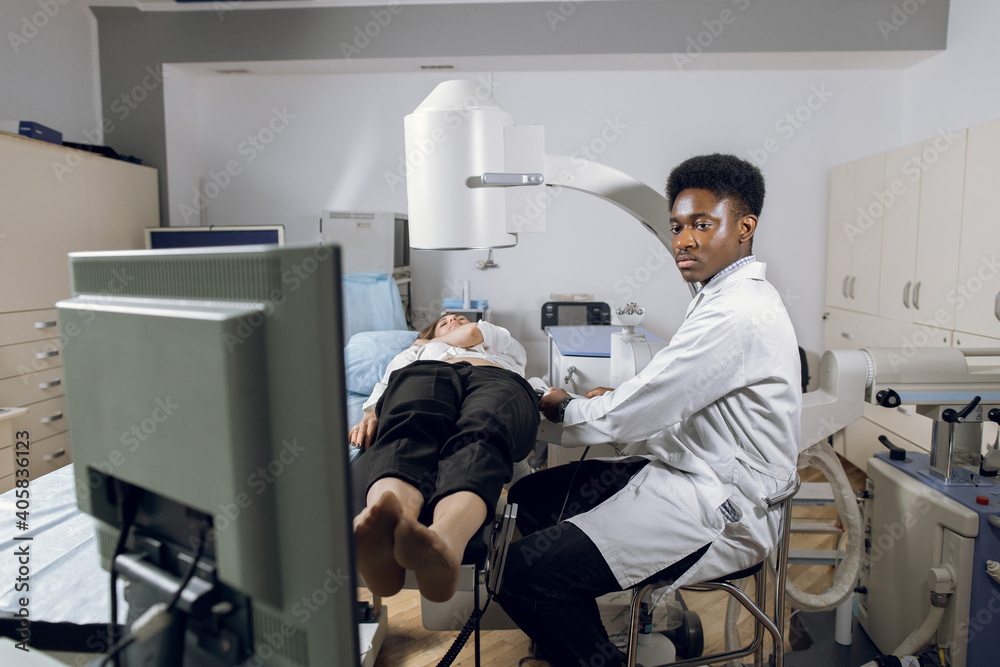 Fototapeta premium Extracorporeal shock wave therapy. Kidney stone treatment. Front view of African male doctor looking at the digital screen of the lithotriptor to determine stones position of woman patient