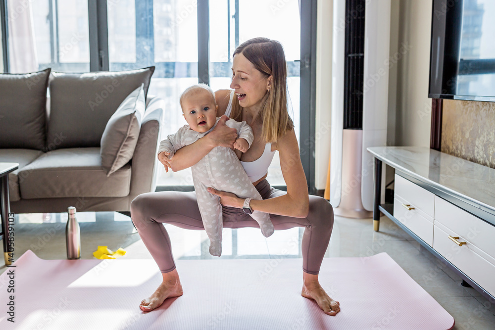 Obraz premium Young mother in sport clothing exercising at home with baby. Online training during coronavirus covid-19 quarantine. Stay fit and safe during pandemic lockdown. Sport, fitness, healthy concept