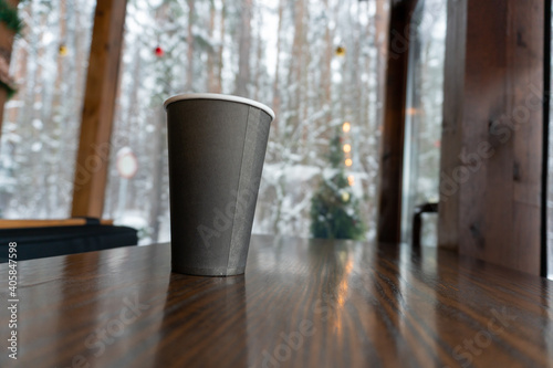 A paper cup of coffee stands on a wooden counter in a pub and a winter forest is visible in the window photo