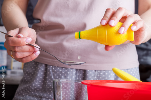 Female hands hold spoon and bottles of ready-made lemon juice in and pour it into the kneading dough from cottage cheese and flour in a red bowl against the background of eggs in a yellow substrate.