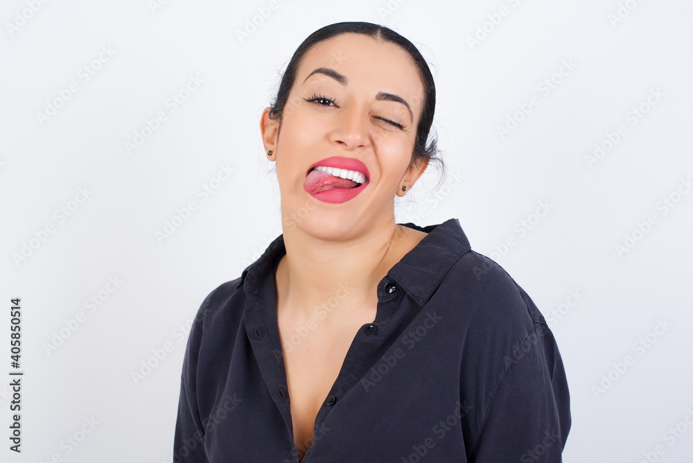 young beautiful Arab woman wearing gray dress against white studio background with happy and funny face smiling and showing tongue. young beautiful Arab woman wearing gray dress against white studio background with happy and funny face smiling and showing tongue.