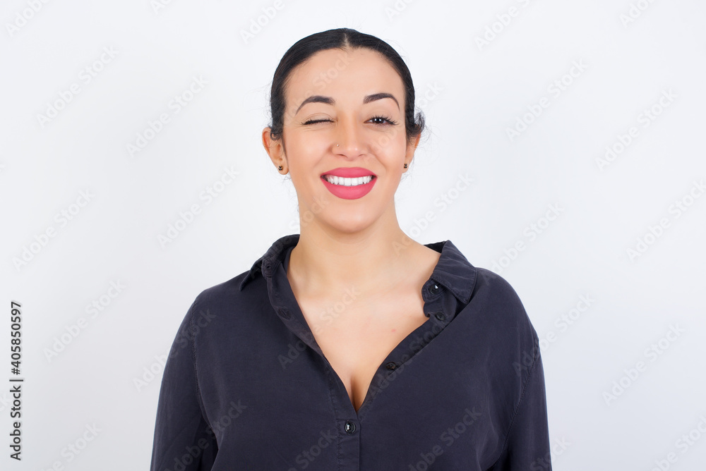 young beautiful Arab woman wearing gray dress against white studio background blinking eyes with pleasure having happy expression. Facial expressions and people emotions concept. young beautiful Arab woman wearing gray dress against white studio background blinking eyes with pleasure having happy expression. Facial expressions and people emotions concept.