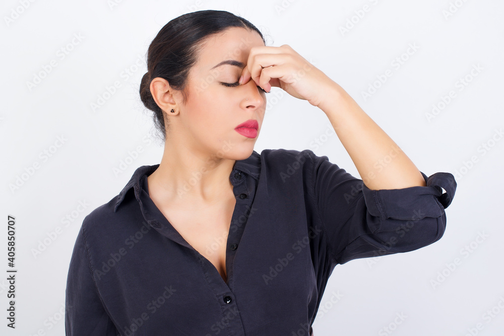 Very upset, young beautiful Arab woman wearing gray dress against white studio background touching nose between closed eyes, wants to cry, having stressful relationship or having troubles with work Very upset, young beautiful Arab woman wearing gray dress against white studio background touching nose between closed eyes, wants to cry, having stressful relationship or having troubles with work