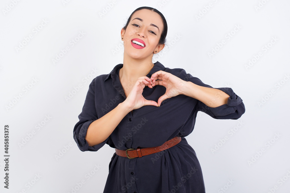 young beautiful Arab woman wearing gray dress against white studio wall smiling in love showing heart symbol and shape with hands. Romantic concept. young beautiful Arab woman wearing gray dress against white studio wall smiling in love showing heart symbol and shape with hands. Romantic concept.