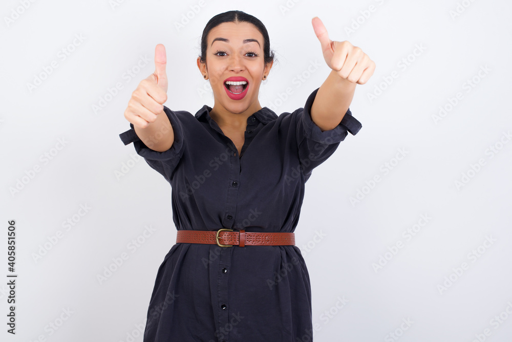 young beautiful Arab woman wearing gray dress against white studio wall making positive gesture with thumbs up smiling and happy for success. Looking at the camera, winner gesture. young beautiful Arab woman wearing gray dress against white studio wall making positive gesture with thumbs up smiling and happy for success. Looking at the camera, winner gesture.